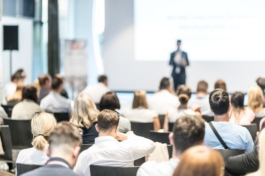 Business and entrepreneurship symposium. Speaker giving a talk at business meeting. Audience in conference hall. Rear view of unrecognized participant in audience.