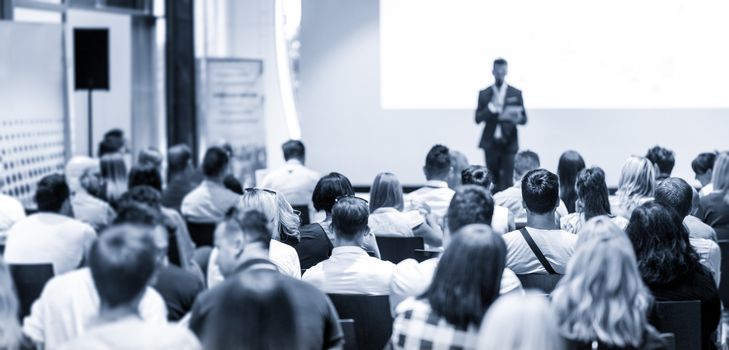 Business and entrepreneurship symposium. Speaker giving a talk at business meeting. Audience in conference hall. Rear view of unrecognized participant in audience.