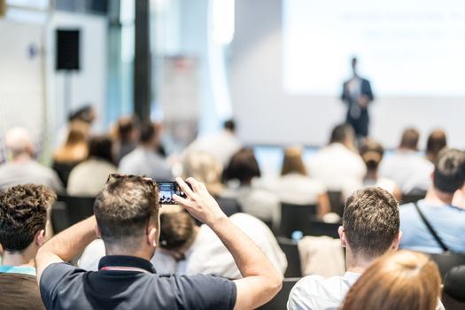 Business and entrepreneurship symposium. Speaker giving a talk at business meeting. Audience in conference hall. Rear view of unrecognized participant in audience taking photo of presentation.