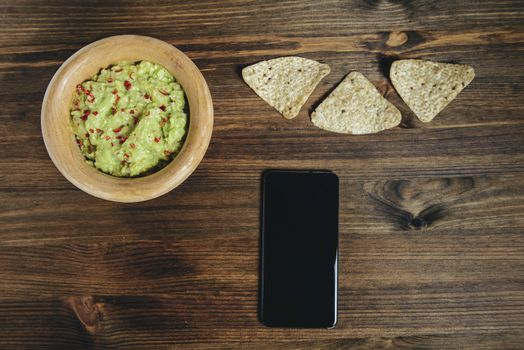 homemade guacamole with nachos and a phone in a wood bowl on a wooden table, typical mexican healthy vegan cuisine, top view and copy space for text