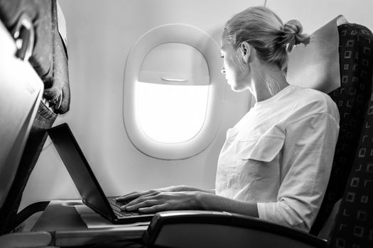 Attractive caucasian female passenger looking through the plain window while working on modern laptop computer using wireless connection on board of commercial flight. Black and white image.