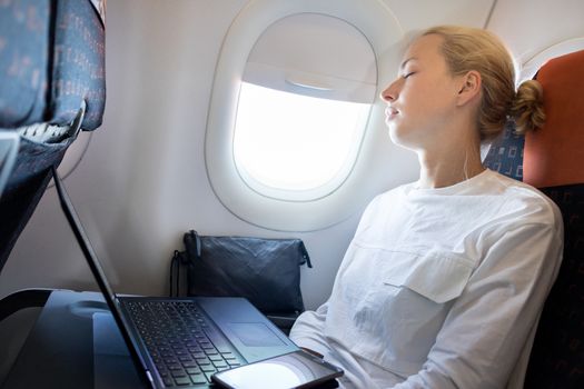 Tired business woman napping on airplane during her business trip woking tasks. Female passenger in flying aircraft sleeps next to the window.