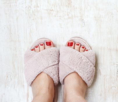 female legs with red nails in home fur fluffy pink slippers on a light wooden background. flat lay. Top view. The concept of a cozy bright girl house.