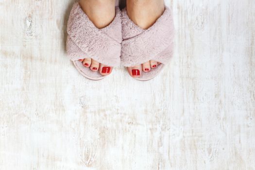 female legs with red nails in home fur fluffy pink slippers on a light wooden background. flat lay. Top view. The concept of a cozy bright girl house.