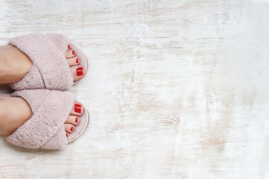 female legs with red nails in home fur fluffy pink slippers on a light wooden background. flat lay. Top view. The concept of a cozy bright girl house.
