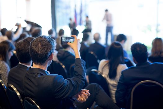 Businessman takes a picture of corporate business presentation at conference hall using smartphone. Business and Entrepreneurship concept. Focus on unrecognizable person in audience.