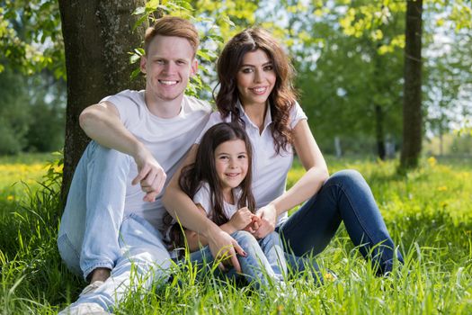 Portrait of happy family sitting on grass under a tree in park on a sunny day