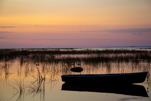 Silhouette of a fishing boat at anchor, reflected in the calm and clear water of the lake, covered with sedge at dusk against the background of a beautiful multi-colored sunset sky.