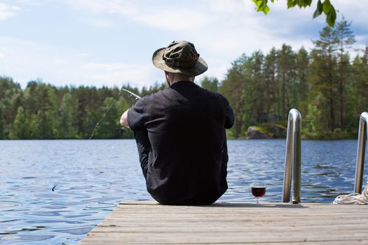 Mature man fishing from wooden pier near cottage on lake in Finland at summer