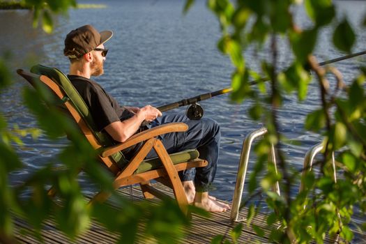 Mature man fishing from wooden pier near cottage on lake in Finland at summer