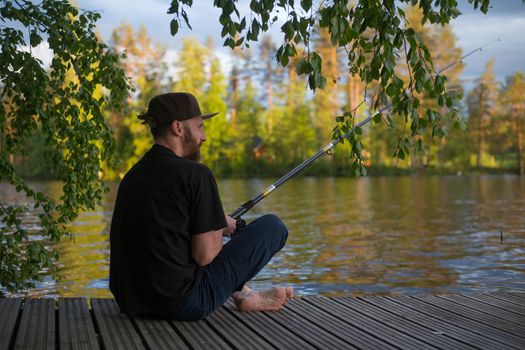 Mature man fishing from wooden pier near cottage on lake in Finland at summer