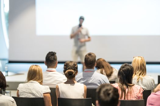 Business and entrepreneurship symposium. Speaker giving a talk at business meeting. Audience in conference hall. Rear view of unrecognized participant in audience.