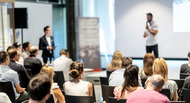 Business and entrepreneurship symposium. Speaker giving a talk at business meeting. Audience in conference hall. Rear view of unrecognized participant in audience.