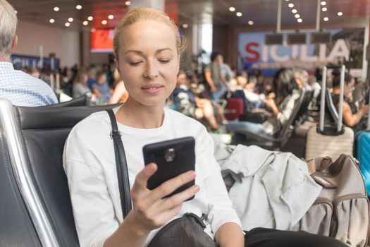 Casual blond young woman reading on her mobile phone while waiting to board a plane at the departure gates at the airport terminal. Travel concept.