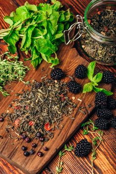 Heap of Dry Green Tea and Fresh Blackberries on Wooden Cutting Board. Bundles of Mint and Thyme Leaves. View from Above.