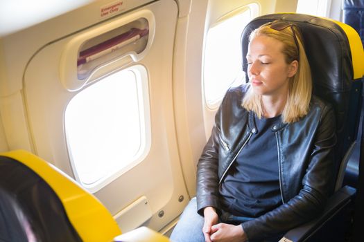 Tired blonde casual caucasian lady napping on exit window seat while traveling by airplane. Commercial transportation by planes.