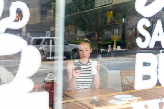 Thoughtful woman reading news on mobile phone during rest in coffee shop. Happy Caucasian female watching her photo on cell telephone while relaxing in cafe during free time.