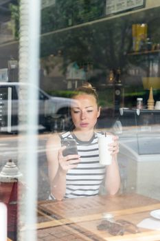 Thoughtful woman reading news on mobile phone during rest in coffee shop. Happy Caucasian female watching her photo on cell telephone while relaxing in cafe during free time.