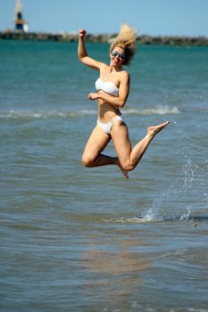 Smiling tanned blonde in a white bikini and in sunglasses makes a jump in the water on the sandy beach of the Baltic Sea shore. sunny day