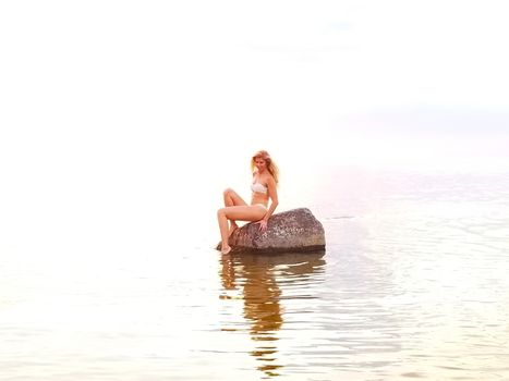 beautiful tanned blonde in a white bikini sits on a stone standing in the water during sunrise