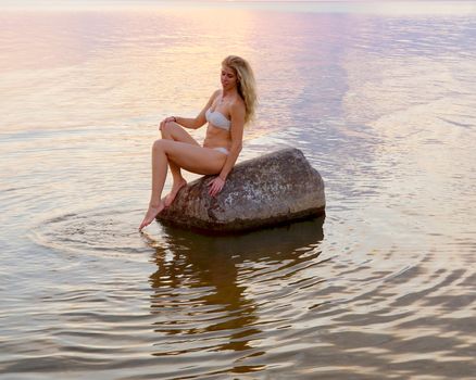 beautiful tanned blonde in a white bikini sits on a stone standing in the water during sunset.