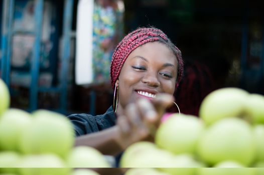 smiling young woman choosing behind  a shelf  an apple  at street market