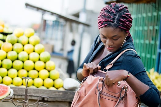 young woman standing in front of a fruit shelf, rummages in her handbag.