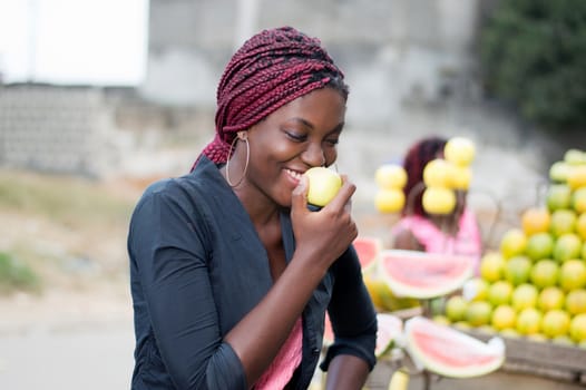smiling young woman standing near a fruit shelf is eating an apple.