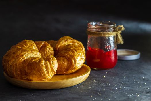 Croissant on wooden dish and strawberry jam bottles in dark background.