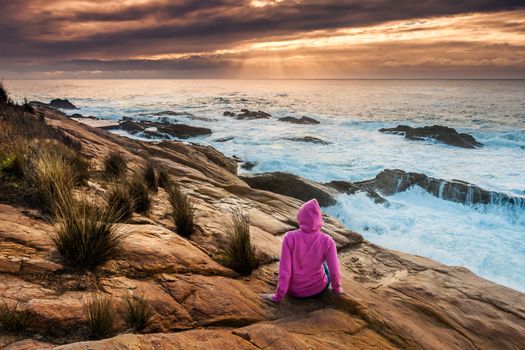 Female in pink hoodie relaxes on coastal rocks watching the sunbeams and sea flows over the craggy rocky coast.
