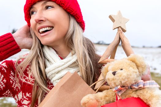 Festive woman in the snowy landscape holding Christmas things, generic teddy bear, shopping bags, and a timber Christmas tree. She is wearing a hand knitted beanie, jumper and scarf