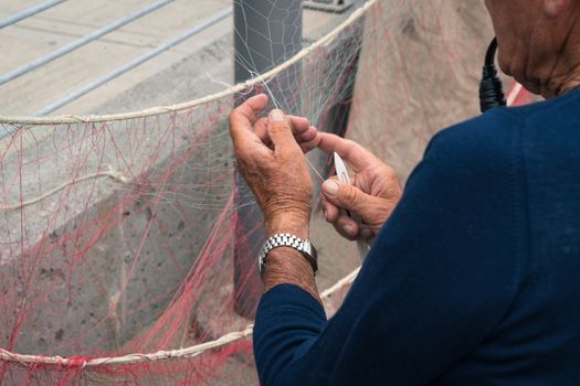 Old fisherman reparing fishing net, sitting with pipe in his mouth.