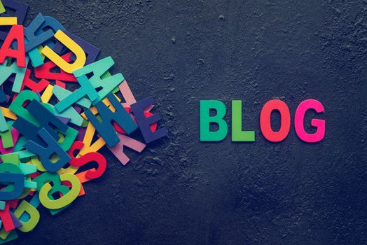 The colorful words "BLOG" made with wooden letters next to a pile of other letters over dark background.