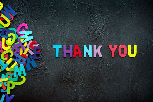 The colorful words "THANK YOU" made with wooden letters next to a pile of other letters over dark background.