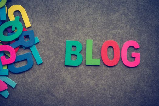 The colorful words "BLOG" made with wooden letters next to a pile of other letters over grey background.