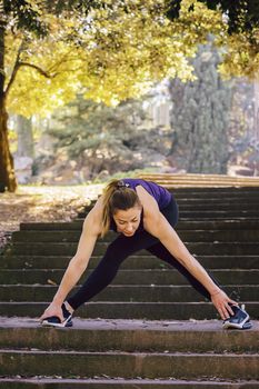 vertical photo of a young sports woman doing some warm up exercises and stretching legs after running, sports girl at the park in morning with sunlight