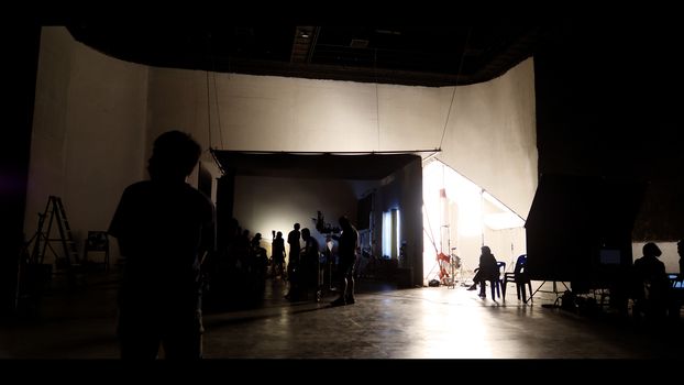 Behind the shooting of video online commercial production and film crew team working and setting light or camera or soft box and equipment set up in big studio in silhouette style