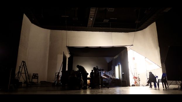 Behind the shooting of video online commercial production and film crew team working and setting light or camera or soft box and equipment set up in big studio in silhouette style