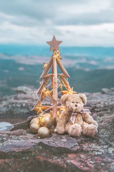 Rustic timber Christmas tree decorated wth golden star lights, gold baubles and a generic brown teddy bear set against a backdrop of the Blue Mountains, Australia