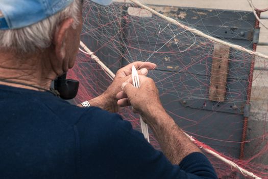 Old fisherman reparing fishing net, sitting with pipe in his mouth.