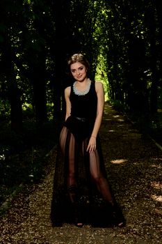 Young attractive woman in the sexy see-through, transparent, transpicuous, diaphanous, clear black dress posing outdoor at the green alley in the old park. Fashion woman. Young woman modern portrait.
