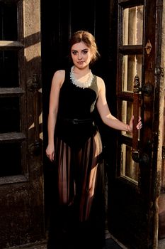Young attractive woman in the sexy see-through, transparent, transpicuous, diaphanous black dress posing at summer sunny day outdoor at the old double door. Fashion woman. Young woman modern portrait.