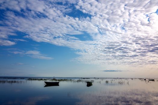 Small fishing boats at anchor, reflected in the calm and clear water of the lake, are covered with sedge in the early morning against the beautiful dawn sky of gentle pastel tones.