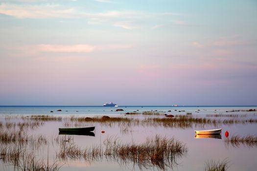 Small fishing boats at anchor, reflected in the calm and clear water of the lake, are covered with sedge in the early morning against the beautiful dawn sky of gentle pastel tones.