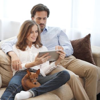 Happy couple using digital tablet at home sitting together with dog on sofa