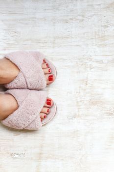 female legs with red nails in home fur fluffy pink slippers on a light wooden background. flat lay. Top view. The concept of a cozy bright girl house.