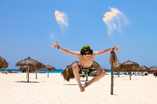A happy young man is jumping for joy on a sand beach background with palm umbrellas, blue sky and ocean. His body and face in the sand and he threw a handful of sand into the sky.