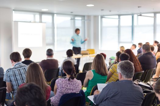 Business and entrepreneurship symposium. Speaker giving a talk at business meeting. Audience in conference hall. Rear view of unrecognized participant in audience.