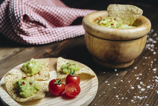 fresh guacamole in a wood bowl with nachos next to a kitchen rag, typical mexican healthy vegan cuisine with rustic dark food photo style