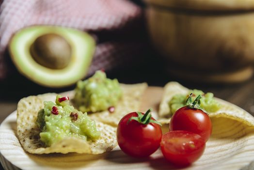 homemade nachos with guacamole in a wood plate to an unfocused avocado and a kitchen rag, typical mexican healthy vegan cuisine with rustic dark food photo style, selective focus